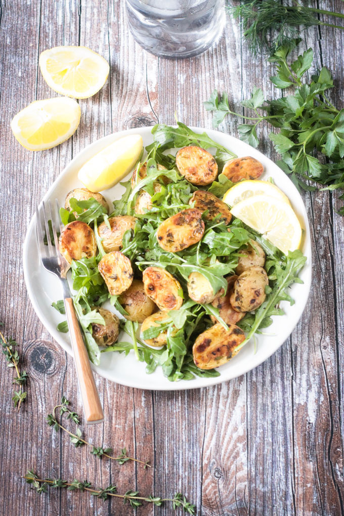 Birds eye image of vegan herbed roasted potato and arugula salad served on a white circular dish on top of a wooden background garnished with fresh lemon slices.