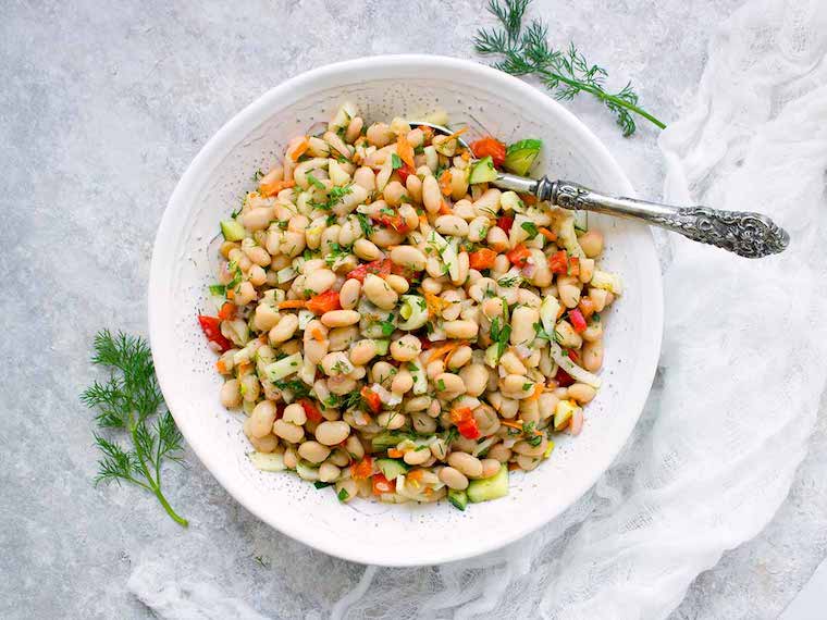 Birds eye image of herbed white bean picnic salad presented in a large white bowl with a silver serving spoon, served alongside fresh herbs on top of a white marble surface