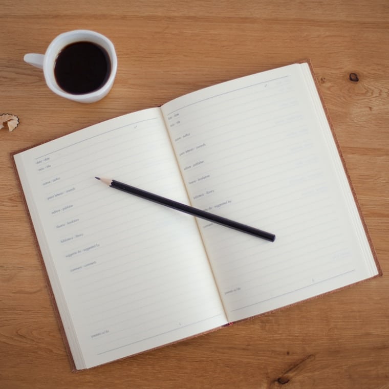 An opened notebook with a pencil on top and a cup of coffee beside it.