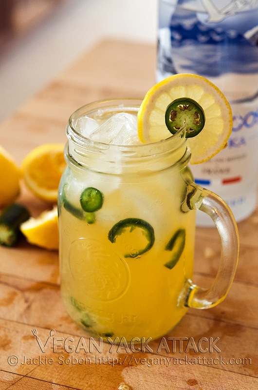 Close up of spicy vegan vodka lemonade presented in a clear large jar, garnished with lemon slices and fresh jalapeno peppers, alongside additional fresh lemon slices in the background 