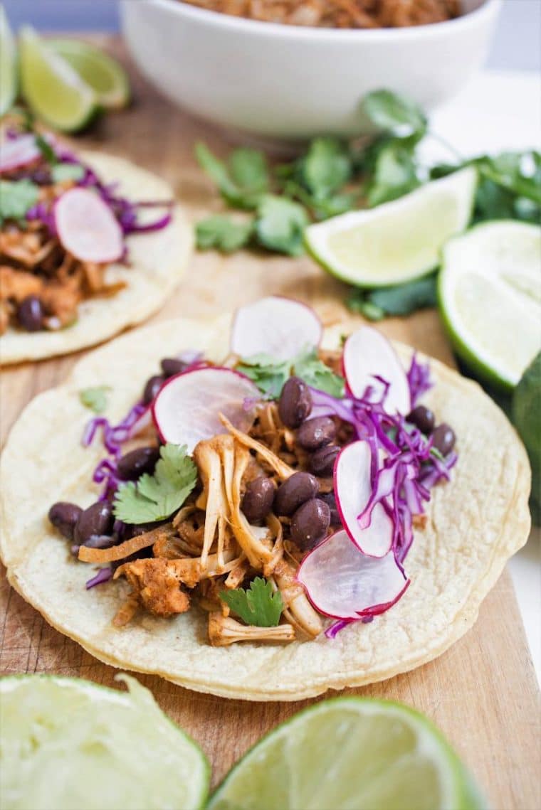 Close up of a plant-based jackfruit carnitas taco in a small tortilla garnished with cilantro, radishes, and cabbage and served with lime slices and extra cilantro leaves on the side