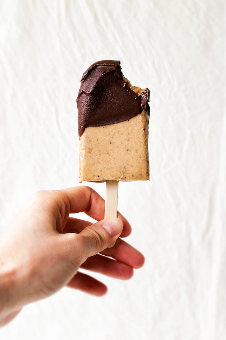 Close up of a hand holding a vegan banana, peanut butter, and chocolate popsicle with a bite in it