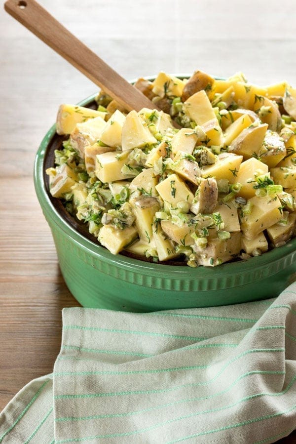 Close up image of plant-based creamy potato salad served in a large green bowl with a wooden serving utensil alongside a green napkin, garnished with additional fresh herbs
