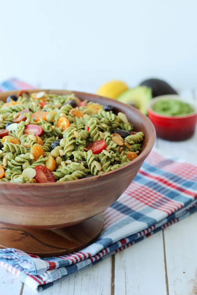 Close up of vegan pesto pasta salad with cherry tomatoes and olives garnished with additional roasted pine nuts served in a large wooden bowl on top of a plaid picnic blanket.