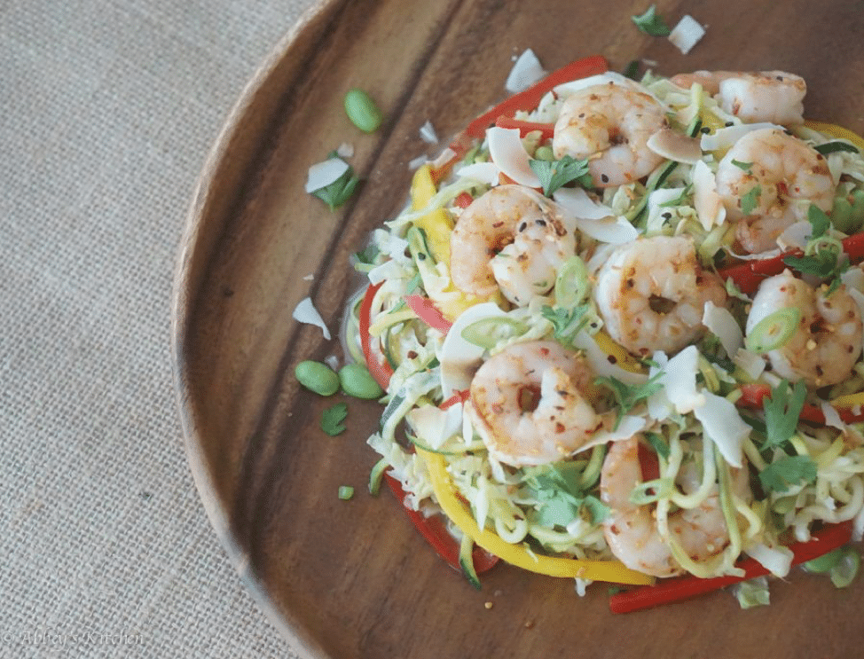 close up image of shrimp, coconut, and zucchini dish served on a wooden plate garnished with herbs and pumpkin seeds