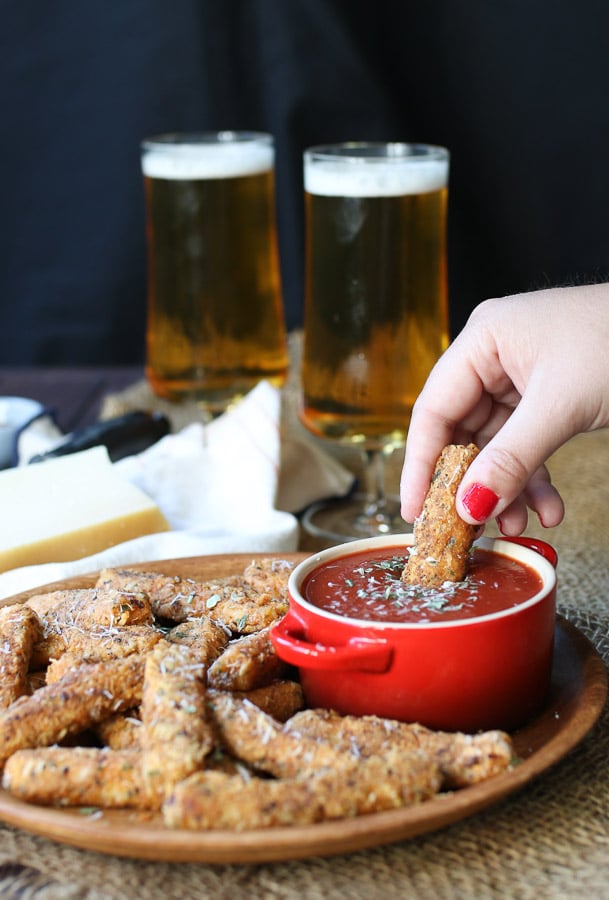 Two fingers dipping a mozzarella stick into marinara sauce in a red ramekin. Two tall glasses of beer in the background.
