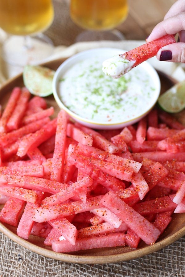 Hand holding a watermelon fry next to dip served in a white bowl, garnished with fresh lime wedges