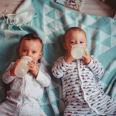 Two babies drinking from bottles while lying on a teal blanket.