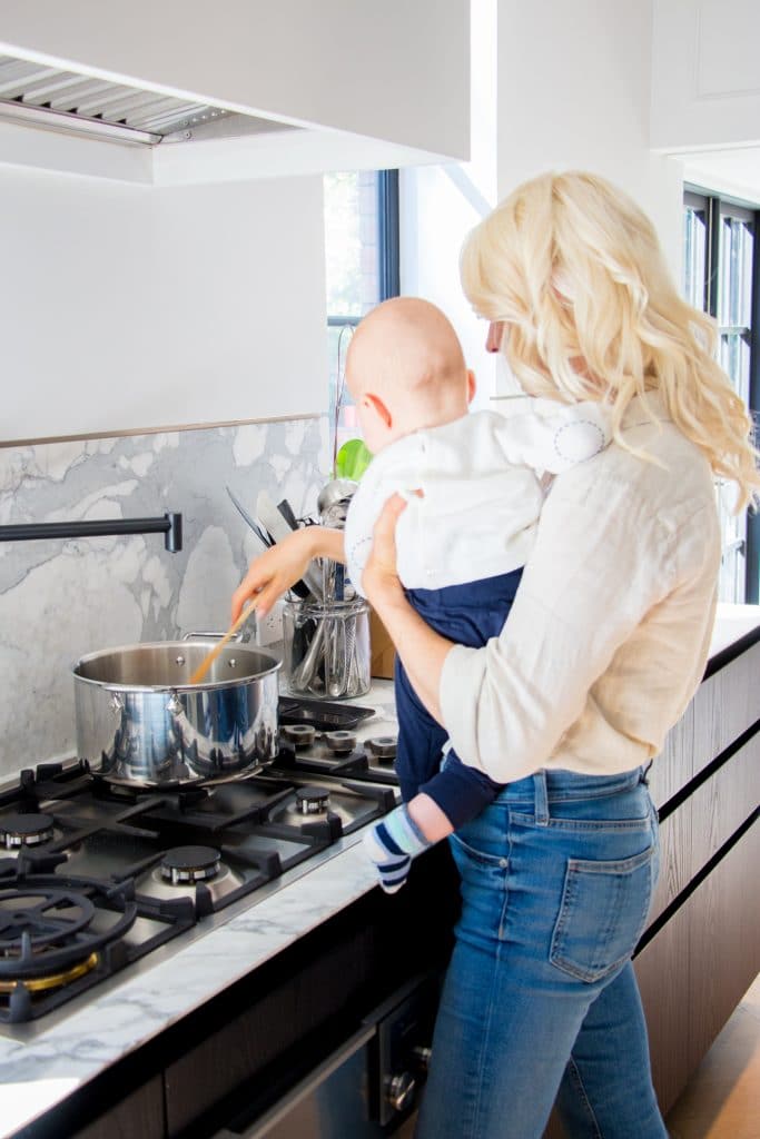 Woman holding her baby next to a stove in their kitchen.