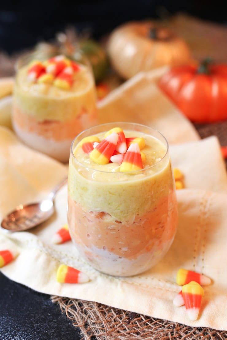 Two clear glasses filled with candy corn flavoured oatmeal topped with candy corn on an orange cloth
