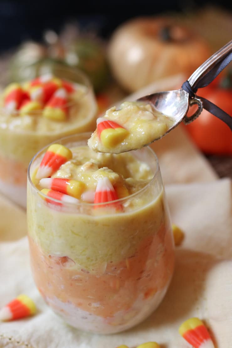 Silver spoon reaching into a clear glass filled with candy corn oatmeal topped with additional candy corn