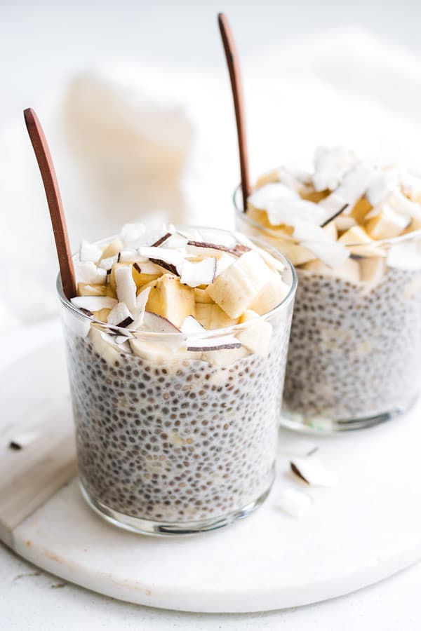 Two glass cups filled with chia pudding and topped with chopped banana and shaved coconut. 