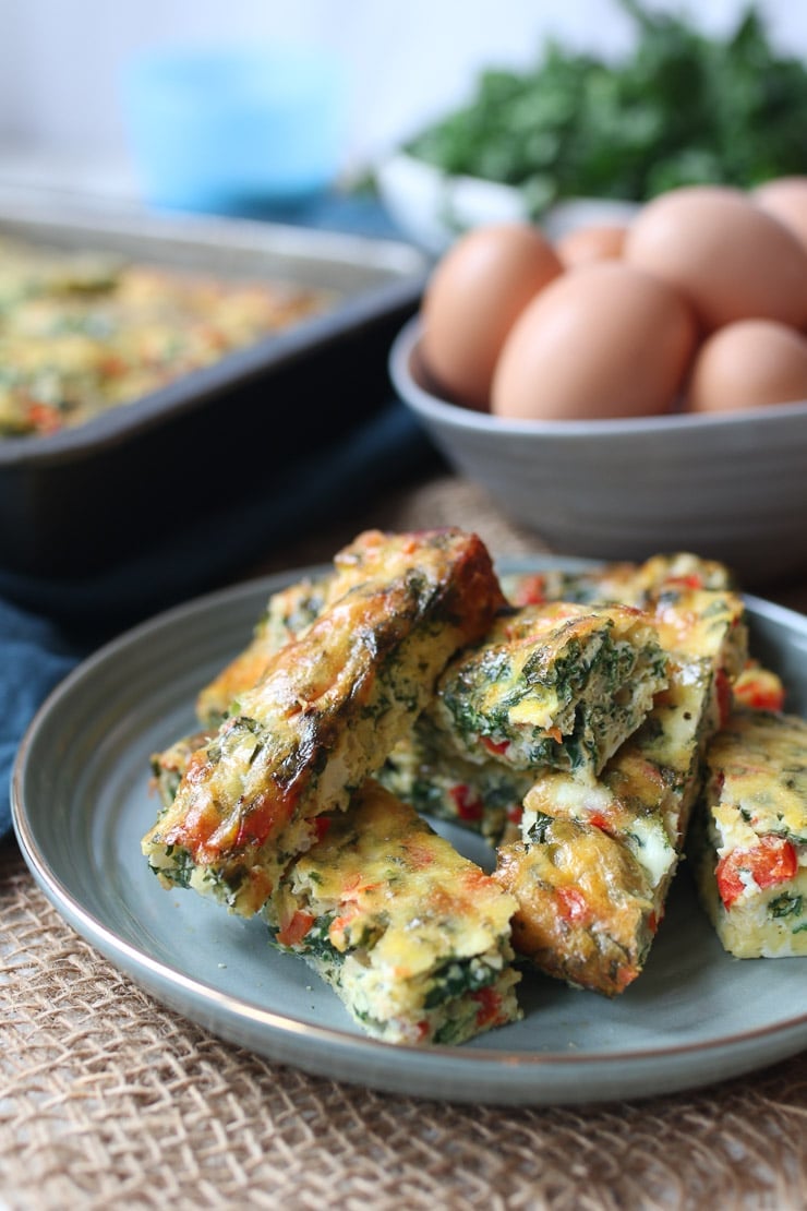 Pile of finger shaped frittata slices on a teal plate with eggs and kale in a bowl in the background.