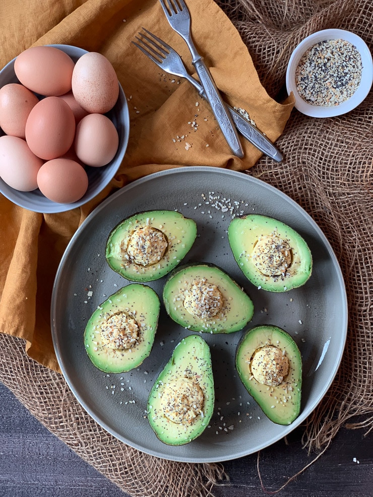 Six avocado halves with egg yolks in the center, sprinkled with everything bagel seasoning. Two forks and a bowl of eggs. 