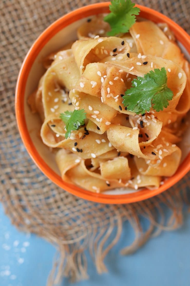 Close up of peanut butter sesame noodles in an orange small bowl topped with black and white sesame seeds and parsley on a blue surface.