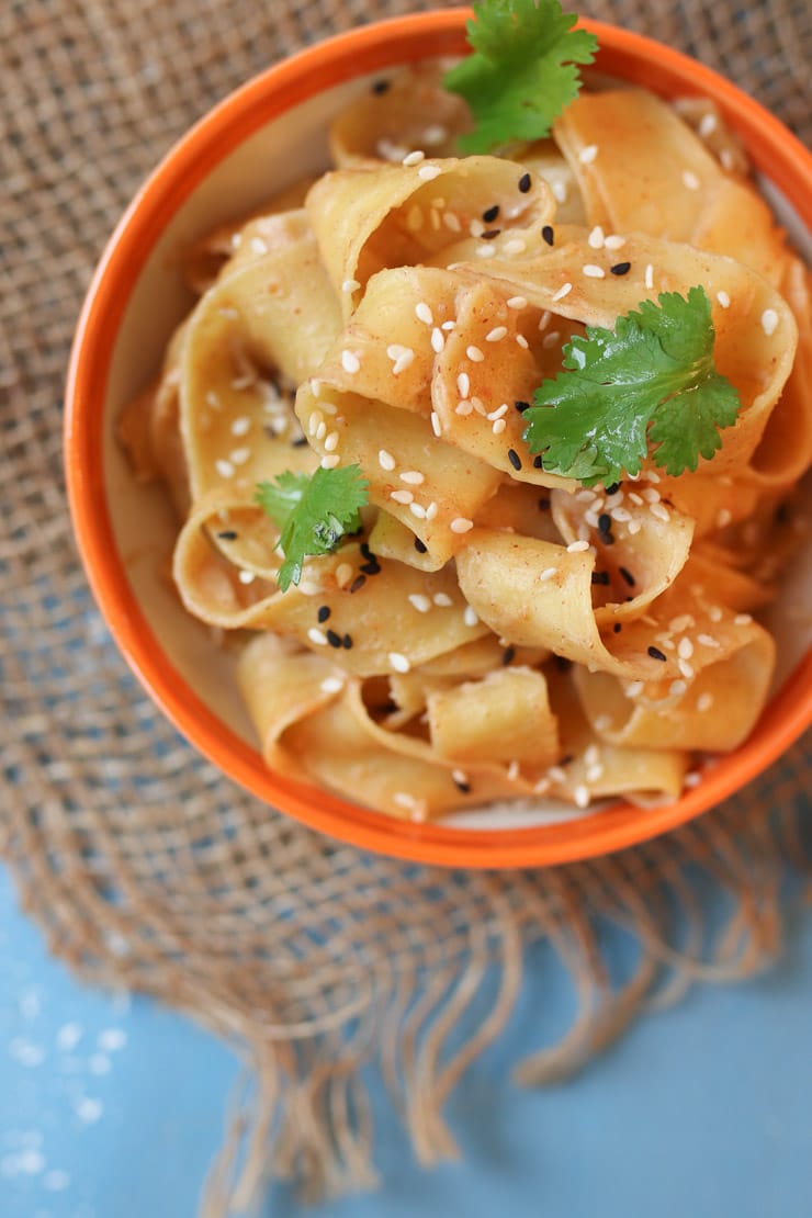 Close up of peanut butter sesame noodles in an orange small bowl topped with black and white sesame seeds and parsley on a blue surface. 