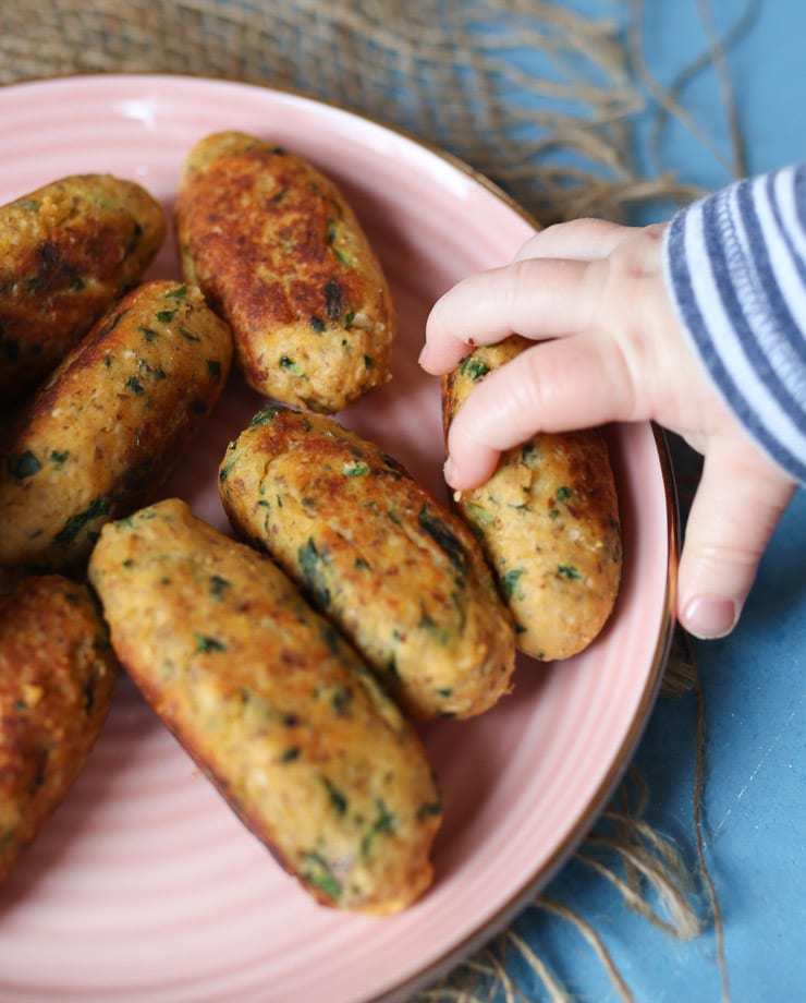 Baby's hand grabbing a finger sized salmon fritter on a rose coloured plate.