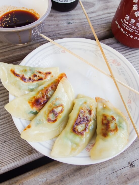 Five potstickers and chopsticks on a white plate with sauce and hot sauce in the background. 