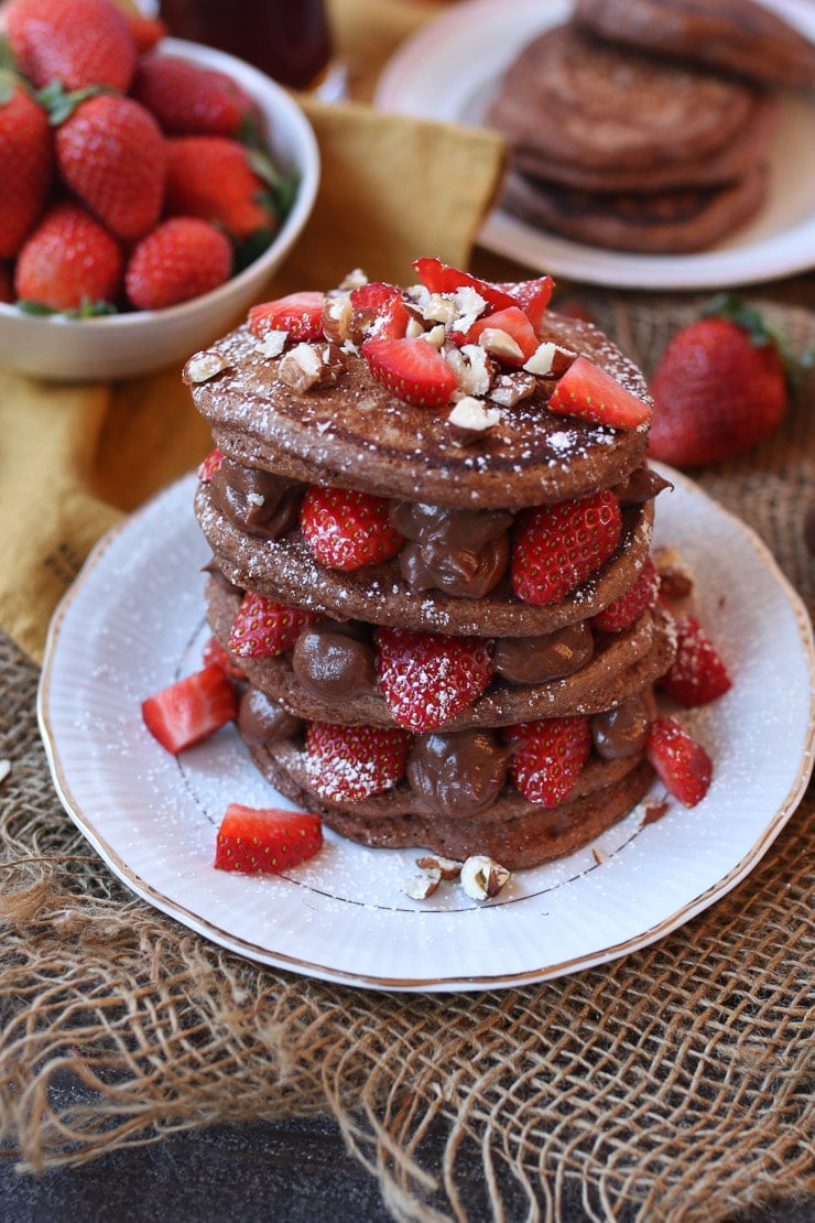 stack of vegan nutella protein pancakes garnished with strawberries and chopped nuts on a white plate