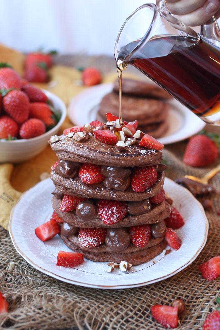 A hand pouring maple syrup over a stack containing four pancakes with a hazelnut chocolate spread in between each layer.