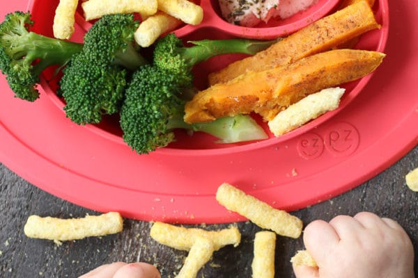 Baby playing with food next to a red plate with baby led weaning lunch.