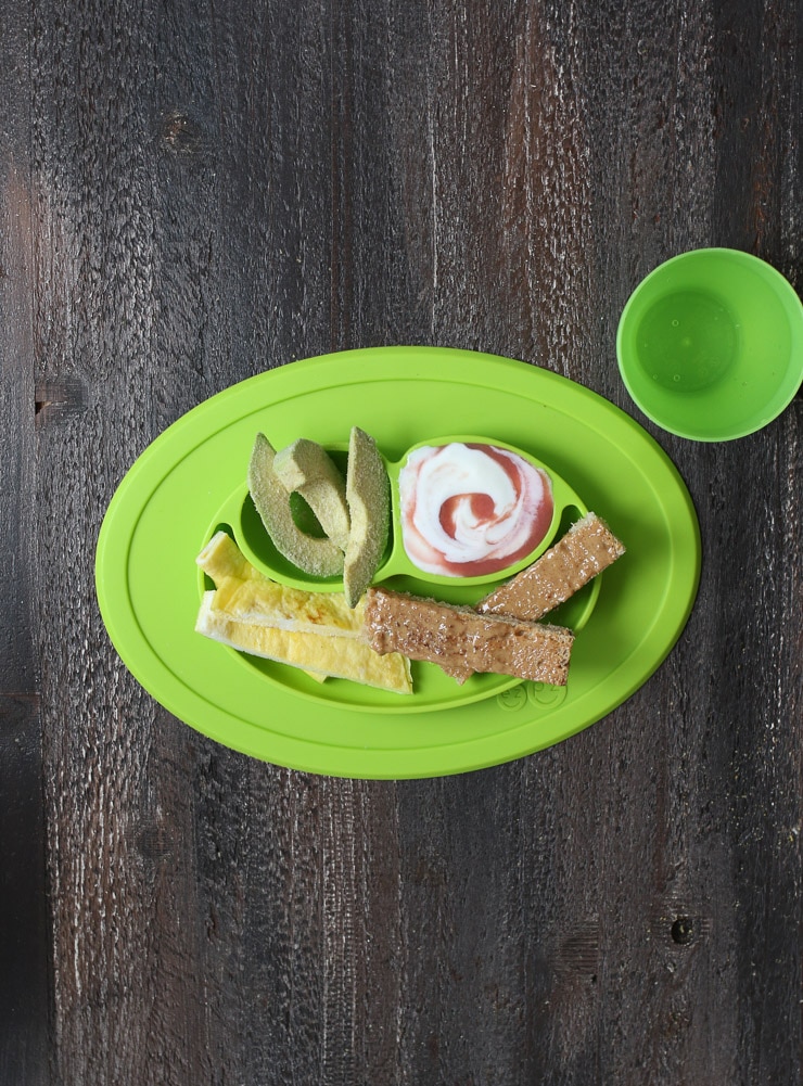 Baby led weaning breakfast containing slices of avocado, omelette strips, strips of toast and yogurt on a green plate 