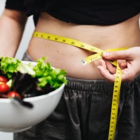 Person measuring their weight circumferance with measuring tape holding a bowl of salad.