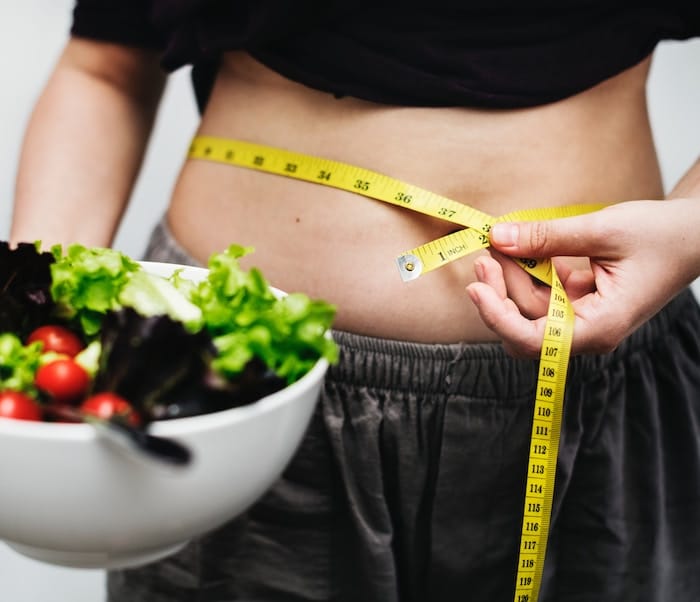 Person measuring their weight circumferance with measuring tape holding a bowl of salad. 