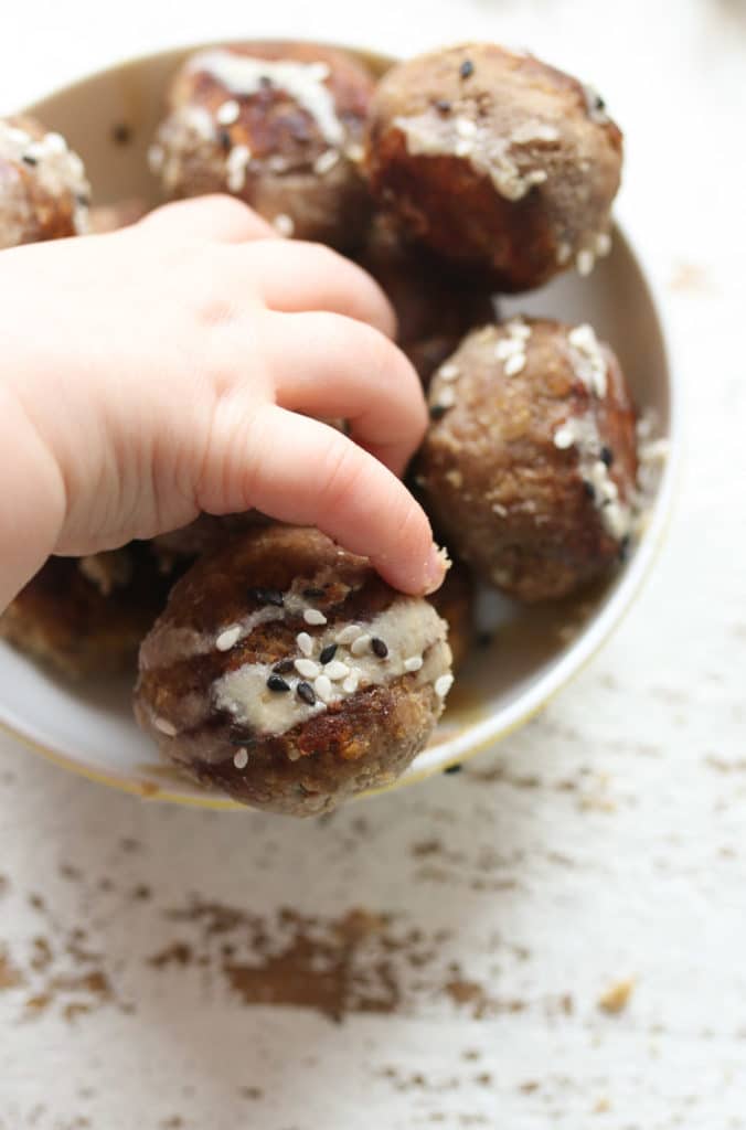 Baby hand reaching for a hummus baby meatball with tahini drizzle and sesame seeds.