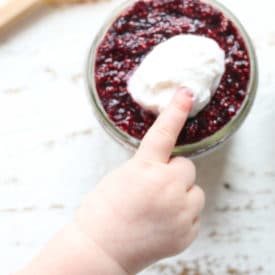 Baby's finger next to vegan chia pudding in a small mason jar.