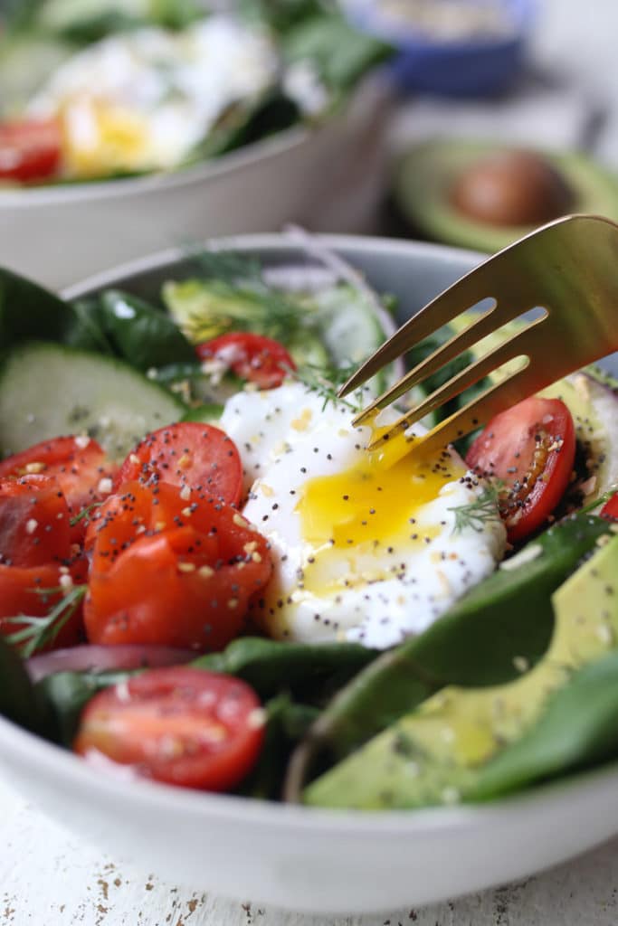 Fork digging into the yolk on a salad with smoked salmon, avocadoes, tomatoes and everything bagel spice.