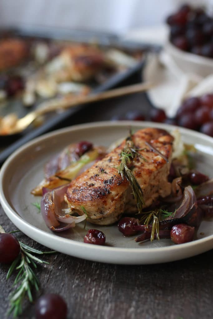 Baked rosemary chicken on a plate with roasted onions and grapes.