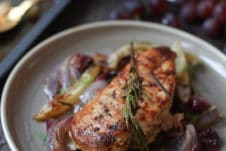 Baked rosemary chicken on a plate with roasted onions and grapes.