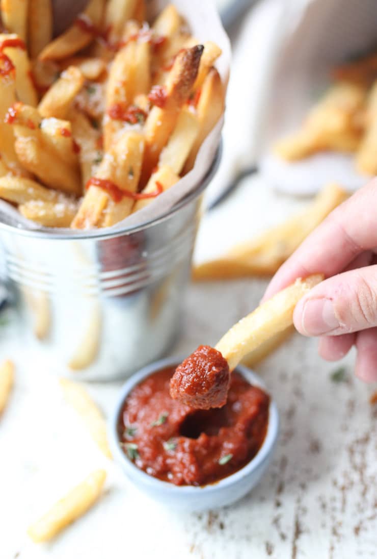 full shot of a hand dipping a french fry into homemade no added sugar ketchup next to a silver bucket containing additional french fries.