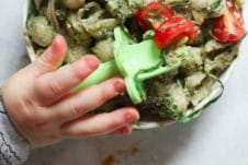 Baby's hand holding a fork over a bowl of avocado pasta.