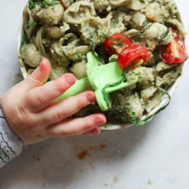 Baby's hand holding a fork over a bowl of avocado pasta.