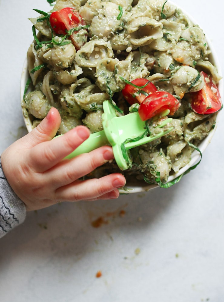 Baby's hand holding a fork over a bowl of avocado pasta.