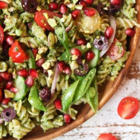 Pistachio parlsey vegan pesto pasta salad on a wooden plate with cherry tomatoes and pomegranates.