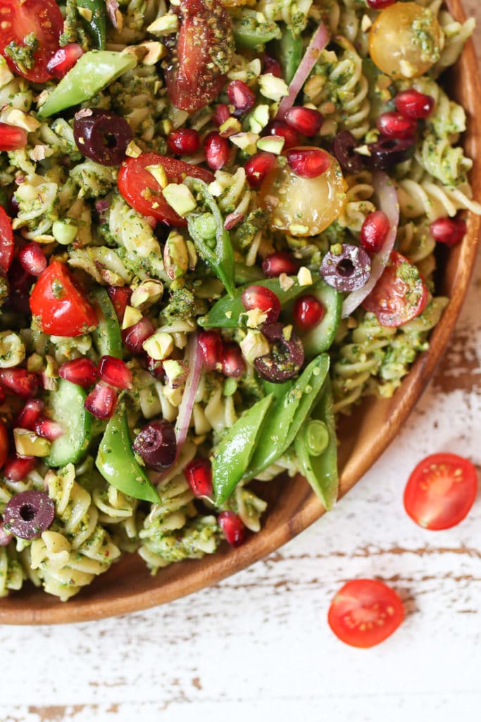 Pistachio parlsey vegan pesto pasta salad on a wooden plate with cherry tomatoes and pomegranates.