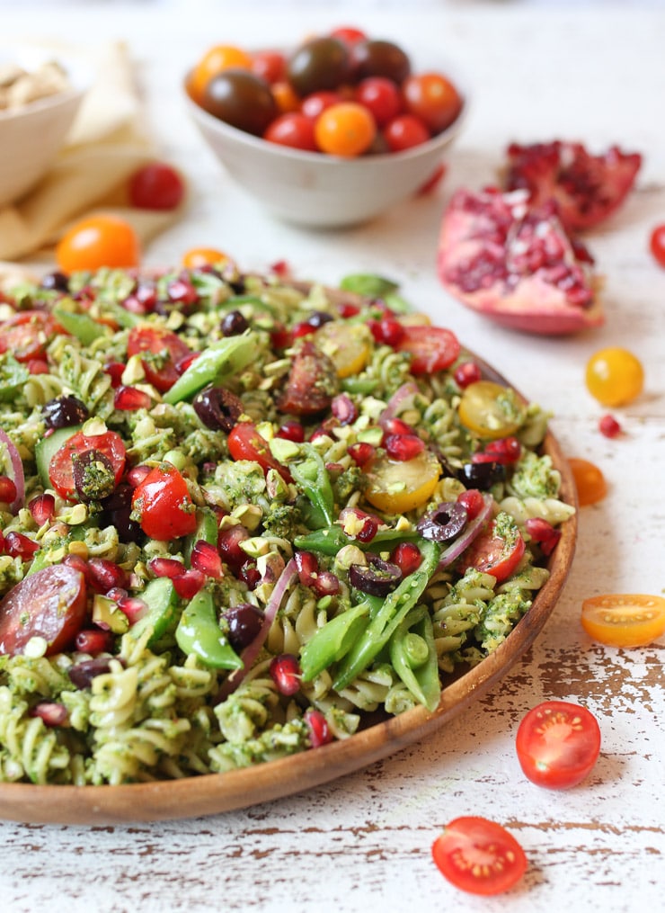 Pistachio parlsey vegan pesto pasta salad on a wooden plate with cherry tomatoes and pomegranates.