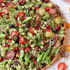 Pistachio parlsey vegan pesto pasta salad on a wooden plate with cherry tomatoes and pomegranates.