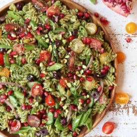 Pistachio parlsey vegan pesto pasta salad on a wooden plate with cherry tomatoes and pomegranates.