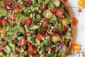 Pistachio parlsey vegan pesto pasta salad on a wooden plate with cherry tomatoes and pomegranates.