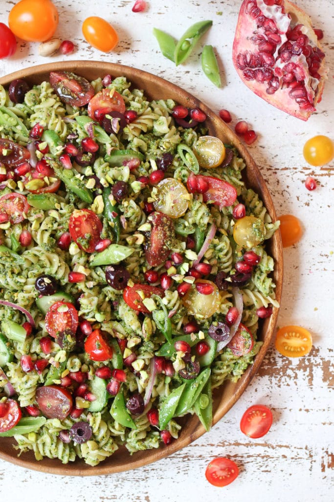 Pistachio parlsey vegan pesto pasta salad on a wooden plate with cherry tomatoes and pomegranates.