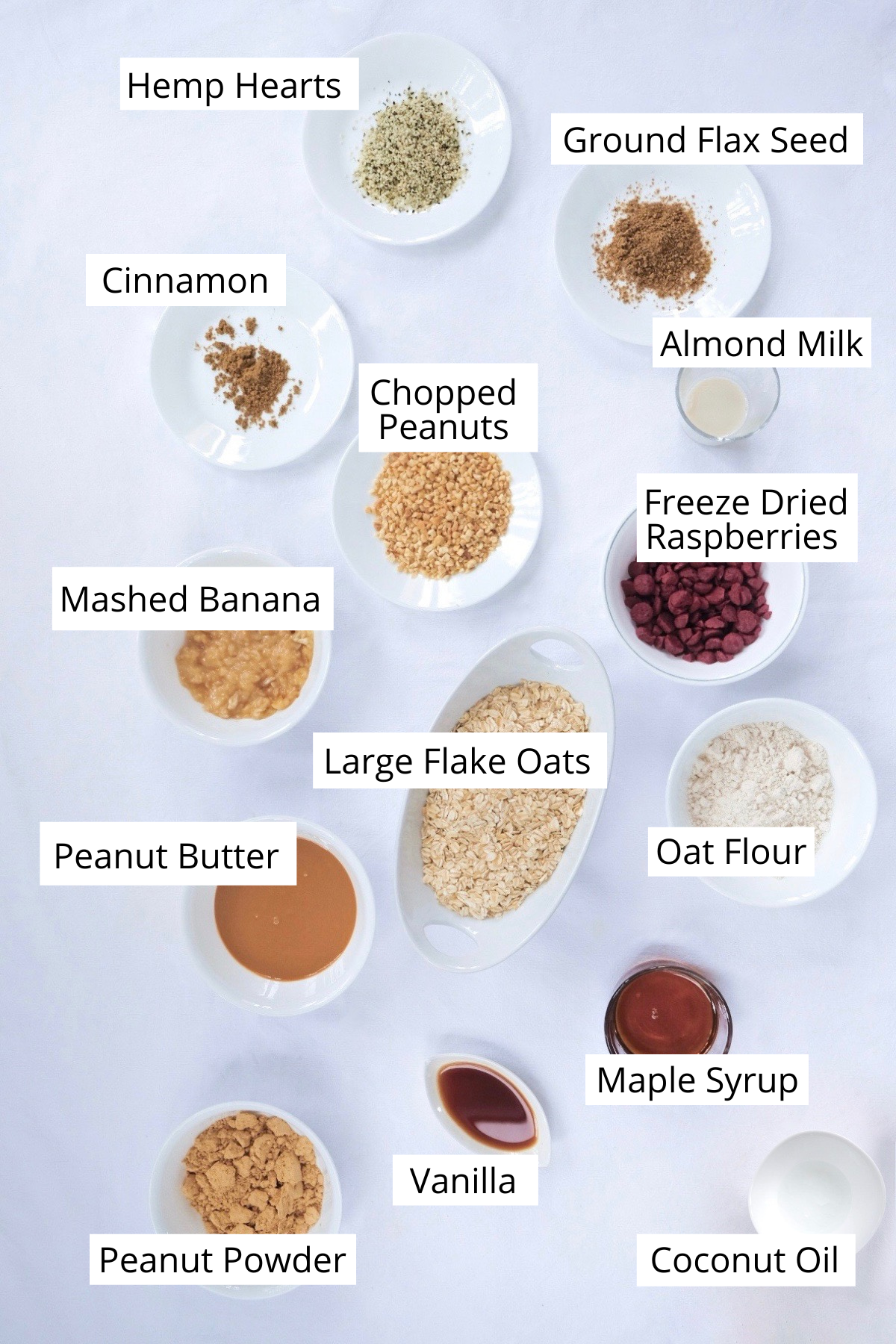 A birds-eye view of the ingredients used to make the peanut butter and jelly cookies, including peanut powder, vanilla, coconut oil, vanilla, maple syrup, oat flour, large flake oats, peanut butter, mashed banana, freeze dried raspberries, chopped peanuts, almond milk, ground flax seed, cinnamon and hemp hearts.