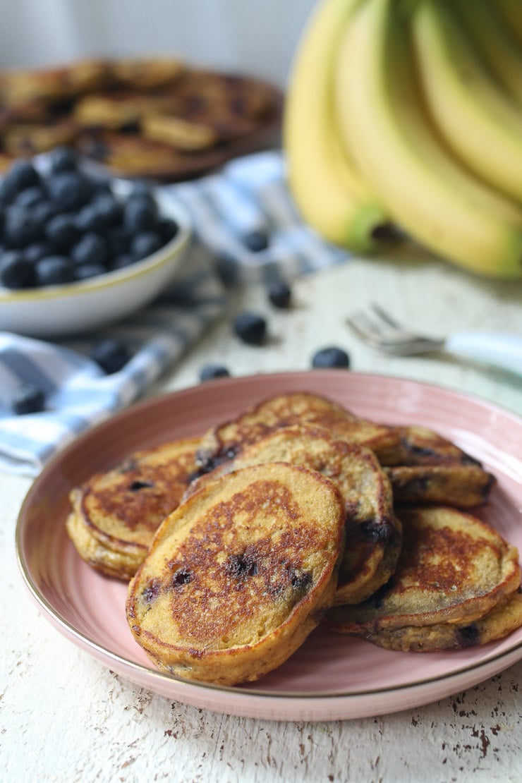 Baby pancakes with blueberries served on a pink plate next to bananas and bowl of blueberries.