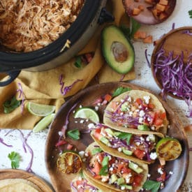 Chicken tacos on a wooden plate next to slow cooker with pulled chicken.