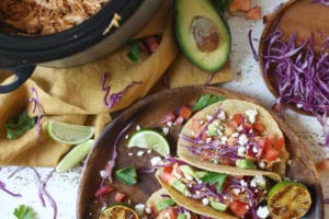 Chicken tacos on a wooden plate next to slow cooker with pulled chicken.