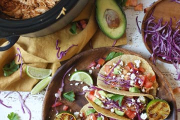 Chicken tacos on a wooden plate next to slow cooker with pulled chicken.