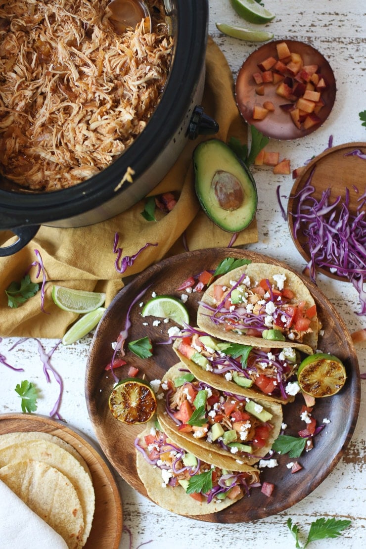 Chicken tacos on a wooden plate next to slow cooker with pulled chicken.